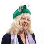Smiling elderly woman wearing a bright green hat with decorative flowers against a white background.