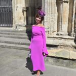 Blog image June - Hats by Sherry Smiling woman in a bright magenta off-shoulder dress with a purple feathered hat posing beside stone columns and steps outside a historic building.