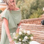 Woman in a pale mint-green dress with a peach fascinator leaning over a round table with a white bouquet outdoors at a garden setting.
