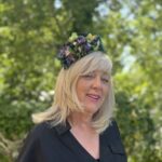 SR Blog Septemebr 26 jpg - Hats by Sherry Smiling blonde woman wearing a colorful flower crown, outdoors in a sunny green garden, wearing a black top.