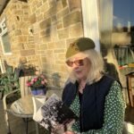 IMG_5493 - Hats by Sherry Older woman with blonde hair and orange sunglasses sits at a glass patio table, reading a black-and-white photo book, wearing a green polka-dot dress and navy quilted vest; a plant pot with pink flowers and a cup are on the table nearby.